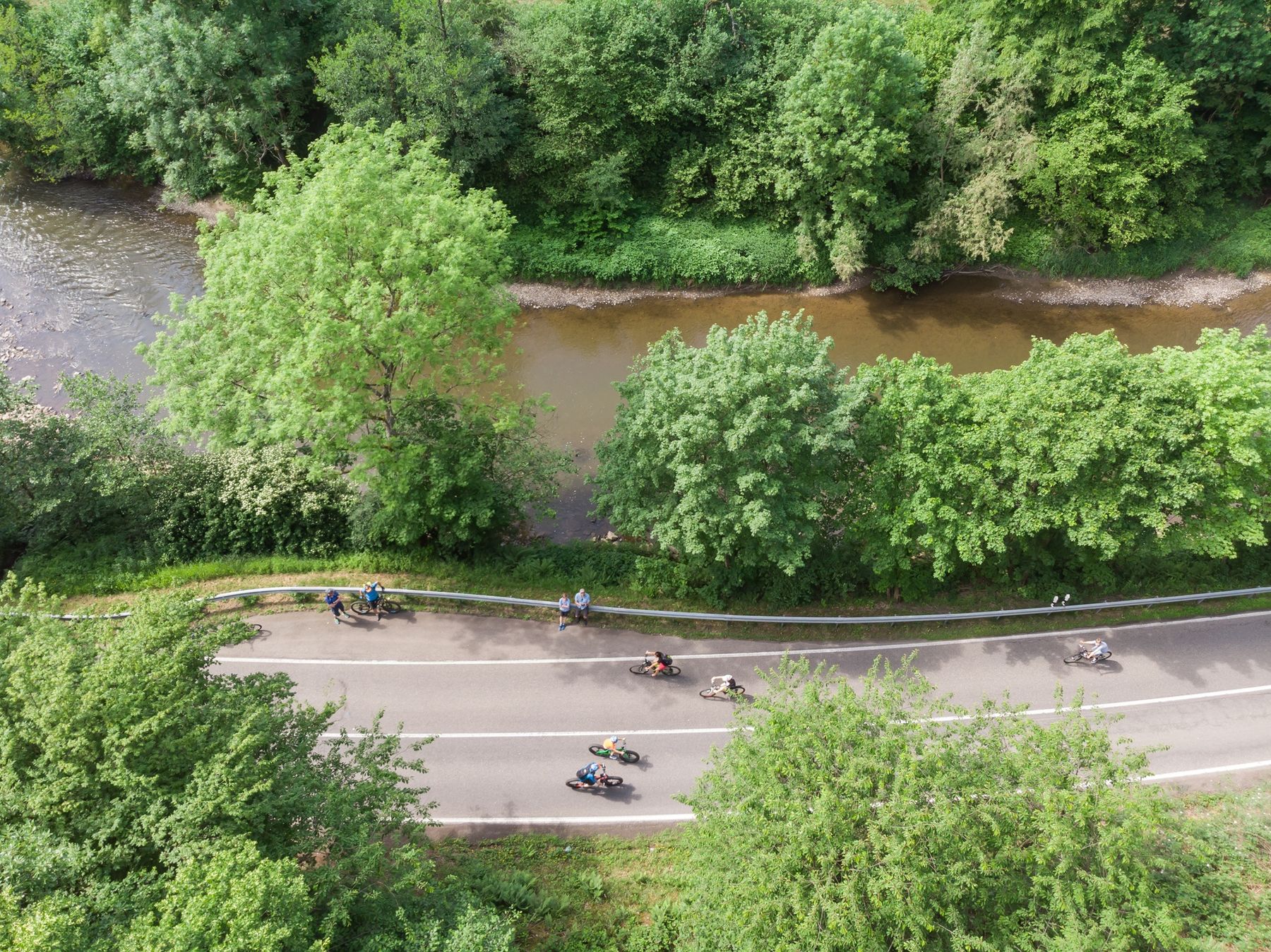 Aerial view of a winding road next to a river, with several motorcyclists riding in a line and lush green trees surrounding the area.